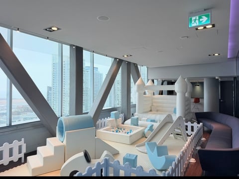 Modern office playroom with blue and white furniture, cityscape views through floor-to-ceiling windows, and wooden support beams