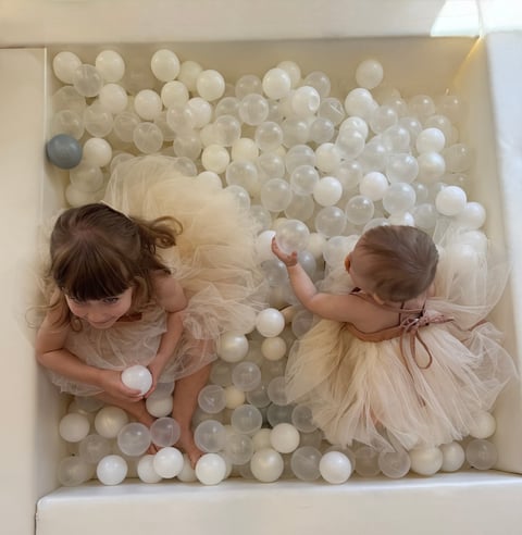 Two toddler girls in tulle dresses playing in a ball pit filled with white and clear plastic balls, viewed from above