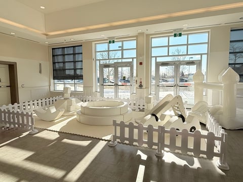 Bright, sunlit indoor space with white picket fencing, sculptural white architectural forms, and large windows overlooking an outdoor area