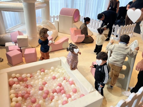 Young children play in an indoor play area with a pink and white ball pit, foam blocks, and climbing structures in a bright, supervised facility